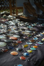 An assortment of colorful stone or gem pendants displayed on a dark cloth background. The pendants are arranged in neat rows and vary in color and pattern, including shades of blue, orange, green, and earth tones. Soft natural light creates a play of shadows, enhancing the visual appeal of the stones.