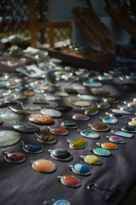 An assortment of colorful stone or gem pendants displayed on a dark cloth background. The pendants are arranged in neat rows and vary in color and pattern, including shades of blue, orange, green, and earth tones. Soft natural light creates a play of shadows, enhancing the visual appeal of the stones.