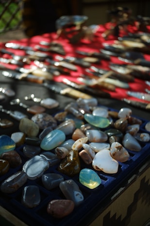 A display of various polished stones and minerals, organized in a grid-like fashion on a dark tray. The background features a red surface with additional miscellaneous objects and subtle shadow patterns.