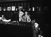 A man in a suit is seated at a large, ornate wooden desk in an office filled with bookshelves and framed pictures. The room is dimly lit, with light focusing on the papers on the desk and the figure at the center.