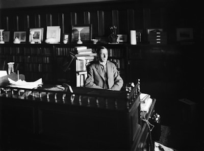 A man in a suit is seated at a large, ornate wooden desk in an office filled with bookshelves and framed pictures. The room is dimly lit, with light focusing on the papers on the desk and the figure at the center.