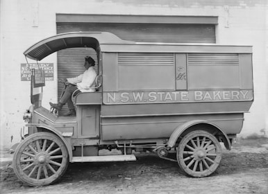 A vintage delivery vehicle labeled 'N.S.W. State Bakery' parked in front of a brick building with a roller door. The vehicle has large wooden wheels and an open cabin where a person is seated in a uniform, appearing relaxed. A sign on the wall says 'Strictly No Admittance Except On Business.'