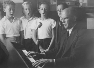 A group of young boys stands near a man playing the piano. The boys have their mouths open, appearing to be singing, while the man is focused on the piano keys. The scene has a vintage, black and white appearance, hinting at an older setting. A microphone is positioned in front of the boys, indicating they might be in a recording or practice session.