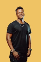 A man with a cheerful expression stands against a solid yellow background. He is wearing a black t-shirt, dark jeans, and several accessories including a watch and bracelets. His posture is relaxed with his hands down by his sides, giving off a friendly and approachable vibe.