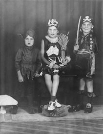 Three children are wearing costumes, each with a distinct theme. The child on the left is dressed in a devil costume with a pointed hat. The middle child is wearing a birthday-themed costume, complete with a crown and a sash. The child on the right is dressed as a pirate, complete with a hat, eyepatch, and a toy sword.