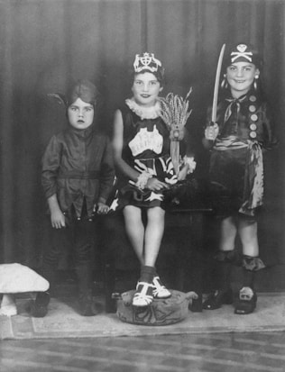 Three children are wearing costumes, each with a distinct theme. The child on the left is dressed in a devil costume with a pointed hat. The middle child is wearing a birthday-themed costume, complete with a crown and a sash. The child on the right is dressed as a pirate, complete with a hat, eyepatch, and a toy sword.