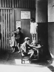 A black and white photograph depicting a group of children in a room. One child is standing on a table being measured, while another sits at a desk with a device. Two more children stand to the side, looking towards the camera. There are signs on the wall related to weight watching.