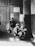 A black and white photograph depicting a group of children in a room. One child is standing on a table being measured, while another sits at a desk with a device. Two more children stand to the side, looking towards the camera. There are signs on the wall related to weight watching.