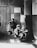 A black and white photograph depicting a group of children in a room. One child is standing on a table being measured, while another sits at a desk with a device. Two more children stand to the side, looking towards the camera. There are signs on the wall related to weight watching.
