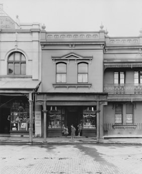 A vintage storefront with ornate architectural details, showcasing a pastry shop with various items displayed in the window. A group of children stands in front of the entrance. The building has large windows, decorative elements, and is flanked by neighboring structures.