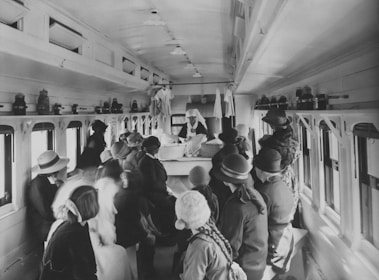 A group of women and children, dressed in early 20th-century attire, gather inside a train car. They appear to be listening to a nurse or healthcare worker standing at a table, wearing a uniform and white cap. The interior of the car includes small windows along both sides, and shelves at the top hold various containers or equipment.