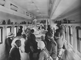 A group of women and children, dressed in early 20th-century attire, gather inside a train car. They appear to be listening to a nurse or healthcare worker standing at a table, wearing a uniform and white cap. The interior of the car includes small windows along both sides, and shelves at the top hold various containers or equipment.
