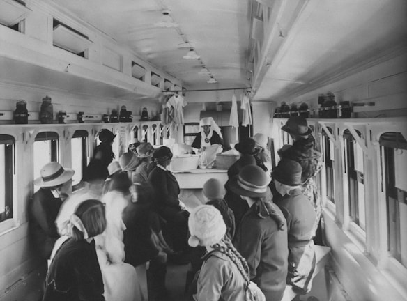 A group of women and children, dressed in early 20th-century attire, gather inside a train car. They appear to be listening to a nurse or healthcare worker standing at a table, wearing a uniform and white cap. The interior of the car includes small windows along both sides, and shelves at the top hold various containers or equipment.