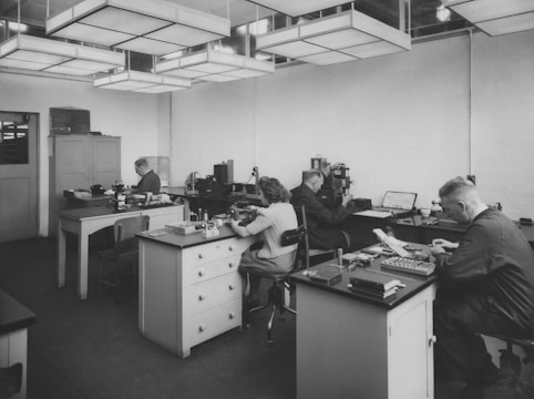 Four people are working at separate desks in an office with various machines, tools, and papers. The room has a sterile and utilitarian feel with rectangular overhead lighting and minimal decorations.
