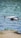 A white drone is captured in mid-flight above a water body, with its camera facing forward and propellers in motion. The background is blurred, showing a mix of blue water and a hint of green vegetation along the shore.