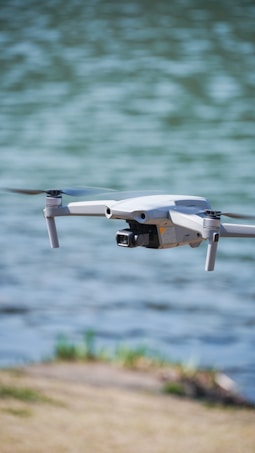 A white drone is captured in mid-flight above a water body, with its camera facing forward and propellers in motion. The background is blurred, showing a mix of blue water and a hint of green vegetation along the shore.
