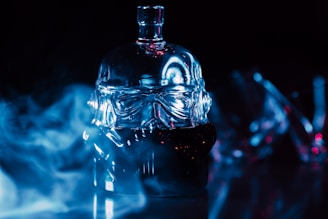 A glass bottle designed in the shape of a helmet is set against a dark background with blue lighting and mist surrounding it. The bottle has intricate detailing and reflects the light, creating a dramatic and mysterious atmosphere.