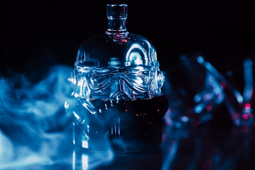 A glass bottle designed in the shape of a helmet is set against a dark background with blue lighting and mist surrounding it. The bottle has intricate detailing and reflects the light, creating a dramatic and mysterious atmosphere.