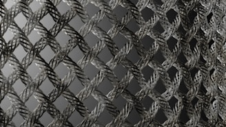A close-up view of an intricately woven metallic mesh pattern with a repetitive, almost geometric design. The strands of metal are interlaced, creating a three-dimensional effect with shadows and highlights. The background is a gradient of dark to light gray, enhancing the depth of the pattern.