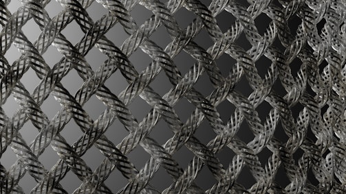A close-up view of an intricately woven metallic mesh pattern with a repetitive, almost geometric design. The strands of metal are interlaced, creating a three-dimensional effect with shadows and highlights. The background is a gradient of dark to light gray, enhancing the depth of the pattern.
