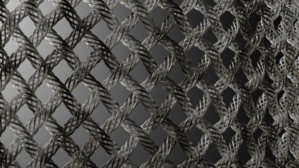 A close-up view of an intricately woven metallic mesh pattern with a repetitive, almost geometric design. The strands of metal are interlaced, creating a three-dimensional effect with shadows and highlights. The background is a gradient of dark to light gray, enhancing the depth of the pattern.