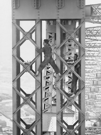 Two men are working high up on a steel structure, likely part of a large bridge or building framework. They are dressed in work clothes and appear to be engaged in construction or maintenance tasks. The background shows the intricate lattice of the structure, indicating an engineering feat.