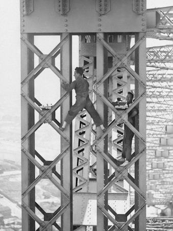 Two men are working high up on a steel structure, likely part of a large bridge or building framework. They are dressed in work clothes and appear to be engaged in construction or maintenance tasks. The background shows the intricate lattice of the structure, indicating an engineering feat.