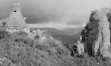 A black and white photograph captures a rugged natural landscape featuring large rock formations surrounded by dense vegetation. In the distance, rolling hills and mountains are visible under an overcast sky. A person stands on a rock outcrop, looking out over the expansive scenery.