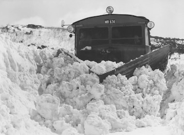 A snowplow is cutting through a large pile of snow, with snowdrifts covering the ground and surrounding landscape. The plow's blade is pushing the snow aside, creating an impressive accumulation in front of it. The area is covered in thick, white snow, and the sky appears overcast.