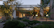 A modern architectural setting with large glass windows reflecting a softly lit interior. The foreground features a lush garden with various trees, shrubs, and rocks. There's a patio area with comfortable seating next to a small water feature, creating a serene outdoor living space.