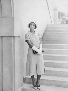 A woman in vintage attire, including a dress, hat, and gloves, stands poised against an architectural structure with stairs. She holds a dark clutch and looks at the camera with a serene expression.