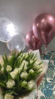 A bouquet of white roses with green leaves is arranged at the bottom. Above, there are several balloons; two are white with golden polka dots, and others are metallic pink, slightly reflecting light. The background has a patterned wall with large green leaves on it.