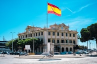 A large, ornate building with classical architecture featuring multiple windows and a clock on its facade. A tall flagpole stands in front of the building, displaying the Spanish flag. Several trees line the street, and there are parked cars and a few signs visible. The sky is clear and bright blue with a single airplane contrail.