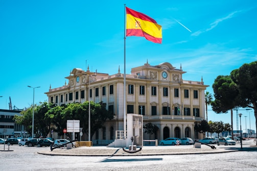A large, ornate building with classical architecture featuring multiple windows and a clock on its facade. A tall flagpole stands in front of the building, displaying the Spanish flag. Several trees line the street, and there are parked cars and a few signs visible. The sky is clear and bright blue with a single airplane contrail.