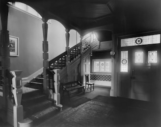 An elegant interior hallway featuring a large, wooden staircase with detailed balusters and handrails. Sunlight streams through stained glass windows on the door and adjacent windows, casting patterns on the floor. The space includes a bench against the wall and framed artwork hanging beside the stairs.