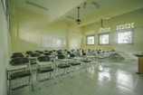 A classroom with rows of empty wooden desks and black chairs, set on a light tiled floor. The room is lit by natural light coming through several windows on one side. In the background, a whiteboard is mounted on the wall. Overhead, there are fluorescent lights and projectors hanging from the ceiling.