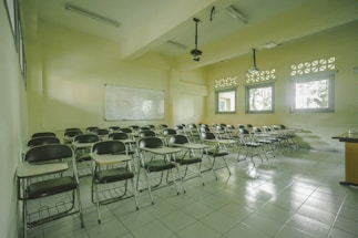 A classroom with rows of empty wooden desks and black chairs, set on a light tiled floor. The room is lit by natural light coming through several windows on one side. In the background, a whiteboard is mounted on the wall. Overhead, there are fluorescent lights and projectors hanging from the ceiling.