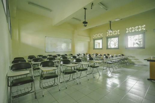 A classroom with rows of empty wooden desks and black chairs, set on a light tiled floor. The room is lit by natural light coming through several windows on one side. In the background, a whiteboard is mounted on the wall. Overhead, there are fluorescent lights and projectors hanging from the ceiling.