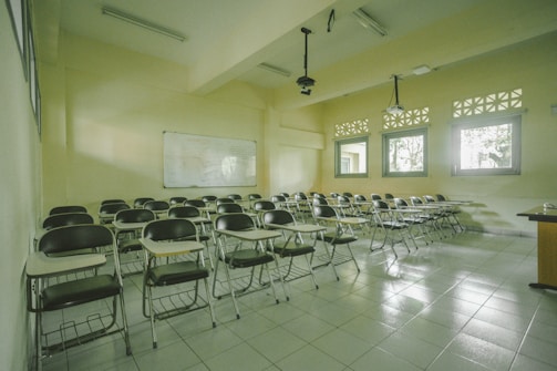 A classroom with rows of empty wooden desks and black chairs, set on a light tiled floor. The room is lit by natural light coming through several windows on one side. In the background, a whiteboard is mounted on the wall. Overhead, there are fluorescent lights and projectors hanging from the ceiling.