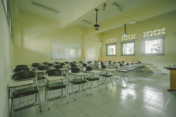 A classroom with rows of empty wooden desks and black chairs, set on a light tiled floor. The room is lit by natural light coming through several windows on one side. In the background, a whiteboard is mounted on the wall. Overhead, there are fluorescent lights and projectors hanging from the ceiling.