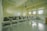 A classroom with rows of empty wooden desks and black chairs, set on a light tiled floor. The room is lit by natural light coming through several windows on one side. In the background, a whiteboard is mounted on the wall. Overhead, there are fluorescent lights and projectors hanging from the ceiling.