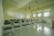 A classroom with rows of empty wooden desks and black chairs, set on a light tiled floor. The room is lit by natural light coming through several windows on one side. In the background, a whiteboard is mounted on the wall. Overhead, there are fluorescent lights and projectors hanging from the ceiling.