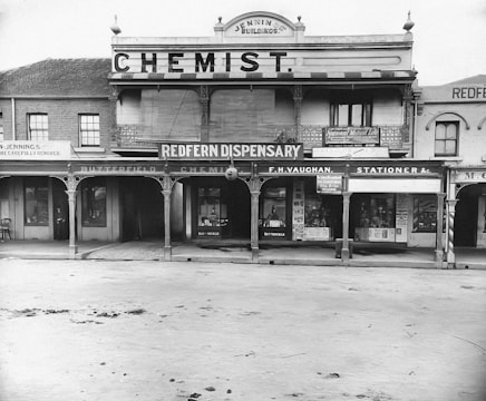 A historic storefront with signage for a chemist and dispensary. The building features architectural details such as columns and arched windows, with various shop names displayed including F.H. Vaughan and a stationer. The scene showcases early 20th-century commercial design.