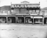A historic storefront with signage for a chemist and dispensary. The building features architectural details such as columns and arched windows, with various shop names displayed including F.H. Vaughan and a stationer. The scene showcases early 20th-century commercial design.