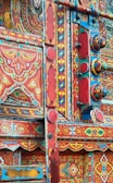 An intricate and colorful display of traditional truck art featuring a complex pattern of geometric shapes and floral motifs. The vibrant design is dominated by reds, blues, and yellows, with decorative elements such as floral designs and painted figures meticulously arranged.