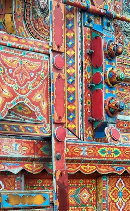 An intricate and colorful display of traditional truck art featuring a complex pattern of geometric shapes and floral motifs. The vibrant design is dominated by reds, blues, and yellows, with decorative elements such as floral designs and painted figures meticulously arranged.