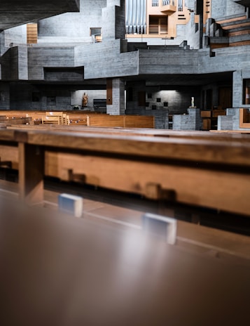A modern and minimalist interior of a concrete building is depicted, featuring geometric and angular architectural elements. The structure is predominantly composed of exposed concrete with wooden seating and panels. A statue and pipe organ add a touch of traditional detail to the otherwise contemporary design.