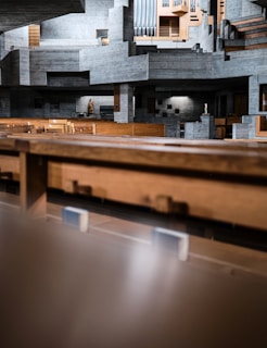 A modern and minimalist interior of a concrete building is depicted, featuring geometric and angular architectural elements. The structure is predominantly composed of exposed concrete with wooden seating and panels. A statue and pipe organ add a touch of traditional detail to the otherwise contemporary design.