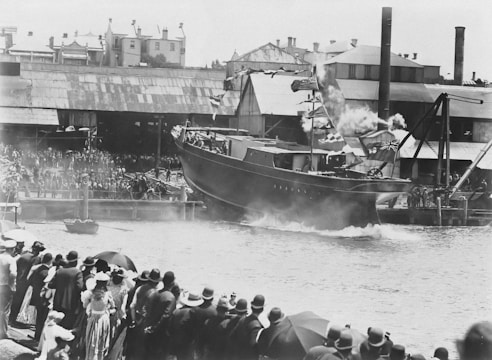 A large crowd gathers near a shipyard to watch the launching of a vessel into the water. The ship is decorated with flags and smoke can be seen coming from its chimney. People are dressed in early 20th-century clothing, including hats and umbrellas. Industrial buildings and a crane are visible in the background, indicating a busy dockside setting.