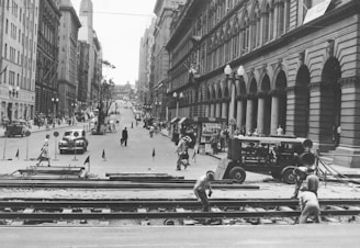 A busy urban street is lined with tall, ornate buildings on both sides. Several pedestrians walk along the sidewalks and cross the street, while a few cars are visible driving down the road. In the foreground, construction workers are actively working on railroad tracks, with some machinery nearby.